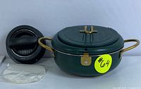 Closed view of the green camping pot with attached lid secured by a brass latch, brass handles on each side, and black burger press with plastic patties mold and press plate laid nearby.