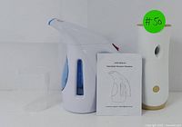 Photo showing handheld garment steamer with water cup and user manual next to the air freshener dispenser with green lot number sticker.