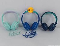 Three headphones shown side by side: light mint green wired on left, medium blue wired in center with cable coiled, dark green wireless on right.