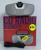 Silver portable CD radio boombox sitting on original red box with 'CD RADIO BOOMBOX' text. A black power cord is coiled and placed in front.