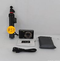 Photo showing compact digital camera, foldable tripod, charging cable, protective pouch, and user manual laid out on a white surface.