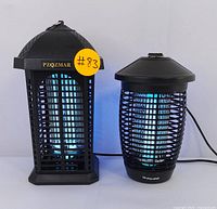 Two electric bug zappers: Pzozmar hexagonal black plastic bug zapper and Phlone cylindrical black bug zapper, both plugged in with blue UV lights active.