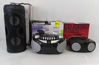 Lot showing all three items: one black cylindrical Bluetooth speaker, one black and silver CD radio boombox, and one black cassette/CD AM/FM radio boombox with original packaging in the background.
