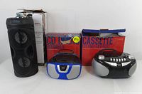 Three Proscan audio devices arranged with their original packaging: a black Bluetooth speaker with dual drivers and circular LED lights, a blue and silver compact CD radio boombox with digital display, and a black and silver cassette/CD AM/FM radio boombox with multiple buttons and dials.