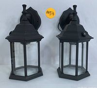 Pair of black outdoor wall lanterns with hexagonal glass panels and top finials seen from front against white backdrop.