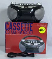 Full front view of Proscan boombox on top of its original red box marked 'CASSETTE CD AM/FM RADIO' with power cord laid out in front.