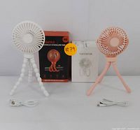 Set of two rechargeable portable fans, one white and one peach-colored, each with flexible tripod legs, USB charging cables, and original product boxes behind them.