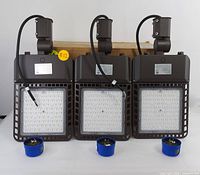 Front view of three brown LED flood lights standing upright with cables and mounting brackets visible. Three blue photocell sensors shown in front of the flood lights.