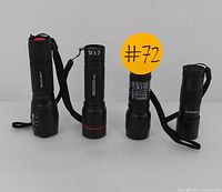 Front view showing four standing black LED tactical flashlights with wrist straps, arranged in a row on a white surface. Labels and branding can be partially seen on the flashlight bodies.