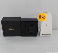 Black Souyie smartwatch displayed in its original black box with gold branding, next to white wireless charging stand.