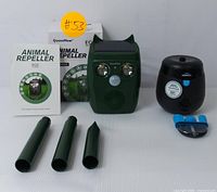 Photo showing the green solar-powered animal repeller with solar panel, two LED lights, a motion sensor, a speaker grille, three detachable green stakes, the black Thermacell mosquito repellent device, and a blue cartridge holder accessory.