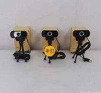Front view of three USB webcams on small black tripods with USB cables coiled next to them, each with its original cardboard box behind.