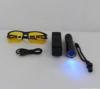 UV flashlight emitting blue light, yellow tinted glasses, USB charging cable, and black carrying case arranged on white background