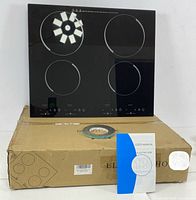 Front view of ceramic hob cooktop on cardboard box, showing four burner circles and touch control panel, with user manual and accessory ring on box.