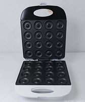 Top and inside view of electric donut maker showing 14 circular donut molds in black non-stick plates, white outer housing, and handle on lid.