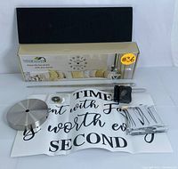 Complete kit items displayed on white background showing clock mechanism, metal clock center, Roman numeral foam pieces, clock hands on acrylic holders, and decorative wall quote sheet, alongside product box showing assembled clock image