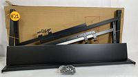 Black adjustable metal keyboard stand parts including two side supports, front panel, assembly hardware, and instruction manual placed against a cardboard background.