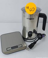 Electric milk frother and digital kitchen scale placed side by side. The frother has a polished stainless steel body with a black handle and the Paris Rhne logo. The kitchen scale is rectangular with a brushed stainless steel surface and digital display.