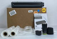 FoodSaver vacuum sealing system with main unit, plastic sealed rolls, accessory pump, connectors, owner's manual, and cardboard box.