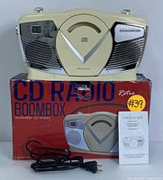 Front view of Proscan retro CD radio boombox showing cream color with silver speaker grills and digital display, placed on top of original red box with power cord and manual visible.