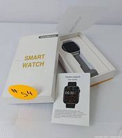 Smartwatch in original white box with label 'Smart Watch', user guide book shown in front, and charging cable inside box.