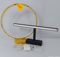 Photo showing modern LED ring lamp with wood-textured circular frame and base, alongside slim rectangular LED desk lamp with black base and USB cables and power adapter.
