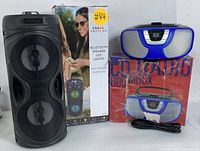 Two audio devices: a black Proscan Bluetooth speaker with LED lights and its box on the left; a blue and gray CD radio boombox with its box and power cord on the right.