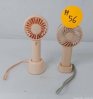 Front view of two peach-colored compact handheld fans standing on their matching charging bases with wrist straps visible, displayed on white background.
