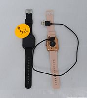 Two smartwatches laid flat on a white surface, one with black band, one with rose band, rose band watch has USB charging cable connected to back.