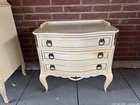 Front view of vintage French Provincial three-drawer nightstand, showing ornate brass ring pulls and cream finish.