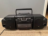 Front view of black JVC portable stereo system with two detachable speakers attached, all placed on a beige carpet against gray wall.