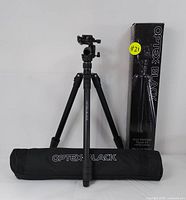 Photo showing the Optex Black tripod fully extended with Arca panhead, carrying case laid in front, and original box standing beside.
