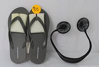 Photo showing gray Amazon Essentials flip-flops with a size sticker and black rechargeable neck fan side by side on a white background.