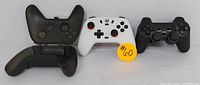 Three items including a black wireless gaming controller, a white wireless gaming controller with orange accents, and a black protective controller case opened.