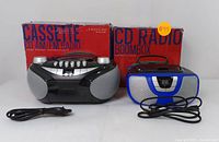 Both Proscan boomboxes shown in front of their original boxes with power cords laid out. Black unit has cassette, CD, and AM/FM radio features; blue unit has CD player and AM/FM radio only.