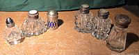 Full lot of six vintage salt and pepper shakers on wooden surface, showing all pieces together with varying glass cuts and metal lids.