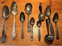 Nine assorted vintage and antique silverplate spoons laid out on wooden surface, showing varying sizes and patterns.