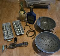 Full view of lot items on wooden floor including metal ice cube trays, colanders, egg beater, horseshoes, and empty bottles