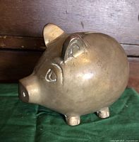 Side and front angled view of a brass piggy bank on a green cloth background showing its shape and facial features.