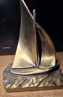 Front view of brass sailboat figurine showing main and secondary sails and base with wave pattern.