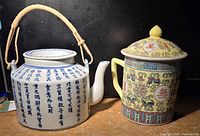 Photo showing both teapots side-by-side on a wooden surface against a dark background. White ceramic teapot with blue characters and woven basket-style handle on the left; yellow and teal hand painted teapot with lid on the right.
