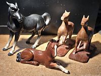 Three horse figurines together: black standing horse, brown and white lying Sylvac horse, and two wooden carved horses with natural wood finish.