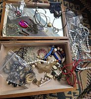 Top view of open wooden jewelry box showing assorted jewelry pieces including chains, bracelets, pendants, and earrings inside the box and some pearl necklaces on the side.