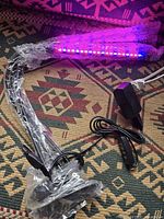 Photo shows an indoor LED grow light with a flexible arm wrapped in plastic, blue and red LEDs lit, attached clip, power plug, and control switch.
