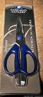 Front view of Chicago Cutlery Instania kitchen scissors in original plastic packaging, showing blue handles and black card backing.
