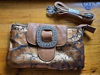 Full view of purse showing distressed gold and black pattern with brown flap and large faux diamond buckle, with unused strap.