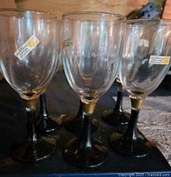 Six clear crystal wine glasses with black stems and gold bands, shown from an angle highlighting the details of the stems and bowls.