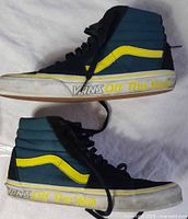 Side view of Vans SK8-Hi sneaker showing black suede and teal upper with yellow side stripe, white sole with 'Vans Off The Wall' text