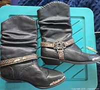 Side profile of both black slouch cowboy boots showing leather upper, ruched shaft, ankle straps with patterned belt and concho buckle, decorative stitching, and metal toe caps.