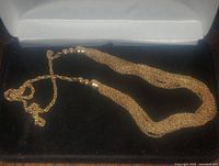 Gold toned multi-strand chain necklace arranged inside a white jewelry box with visible metallic bead accents near the clasp and extending chains.